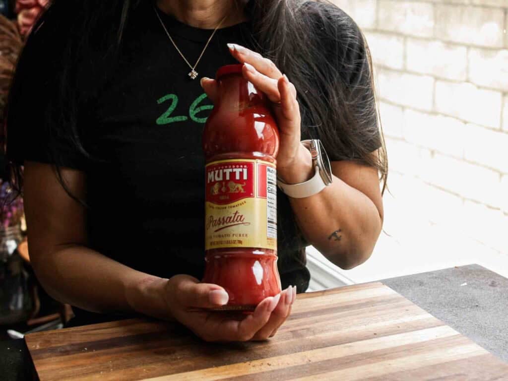 A person holding a bottle of Mutti Passata tomato puree over a wooden cutting board, with part of a kitchen visible in the background.